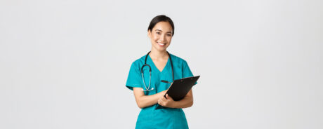 A healthcare professional with a stethoscope around the neck holds a black clipboard and pen, standing against a plain light background.