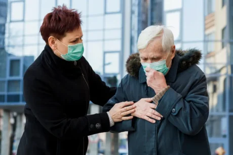 A person in a black coat assisting an elderly individual in a blue coat, both wearing green face masks, outside a modern glass building, showcasing care, support, and health safety during outdoor urban settings.