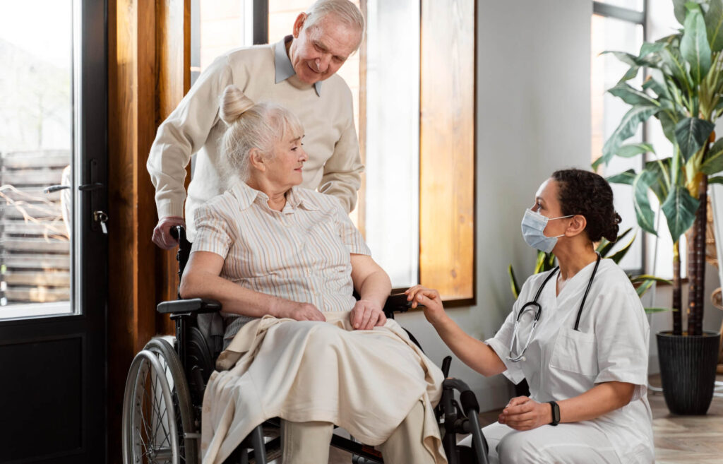 Healthcare professional supporting an elderly woman in a wheelchair while speaking with her and her partner in a UK health and social care setting.