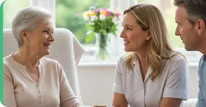 Caregiver in white uniform engaging in supportive conversation with older adults in bright home setting.