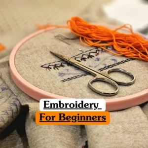 Theoretical textile arts and creative crafting training banner featuring a pink embroidery hoop holding a neutral-coloured fabric with delicate needlework, a pair of silver craft scissors, and a bundle of vibrant orange thread.