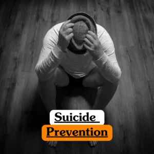 Theoretical mental health awareness and crisis intervention training banner featuring a person in a white long-sleeved shirt sitting on a dark wood floor, head lowered and hands clutching a hat, symbolizing the psychological study of emotional distress and prevention.